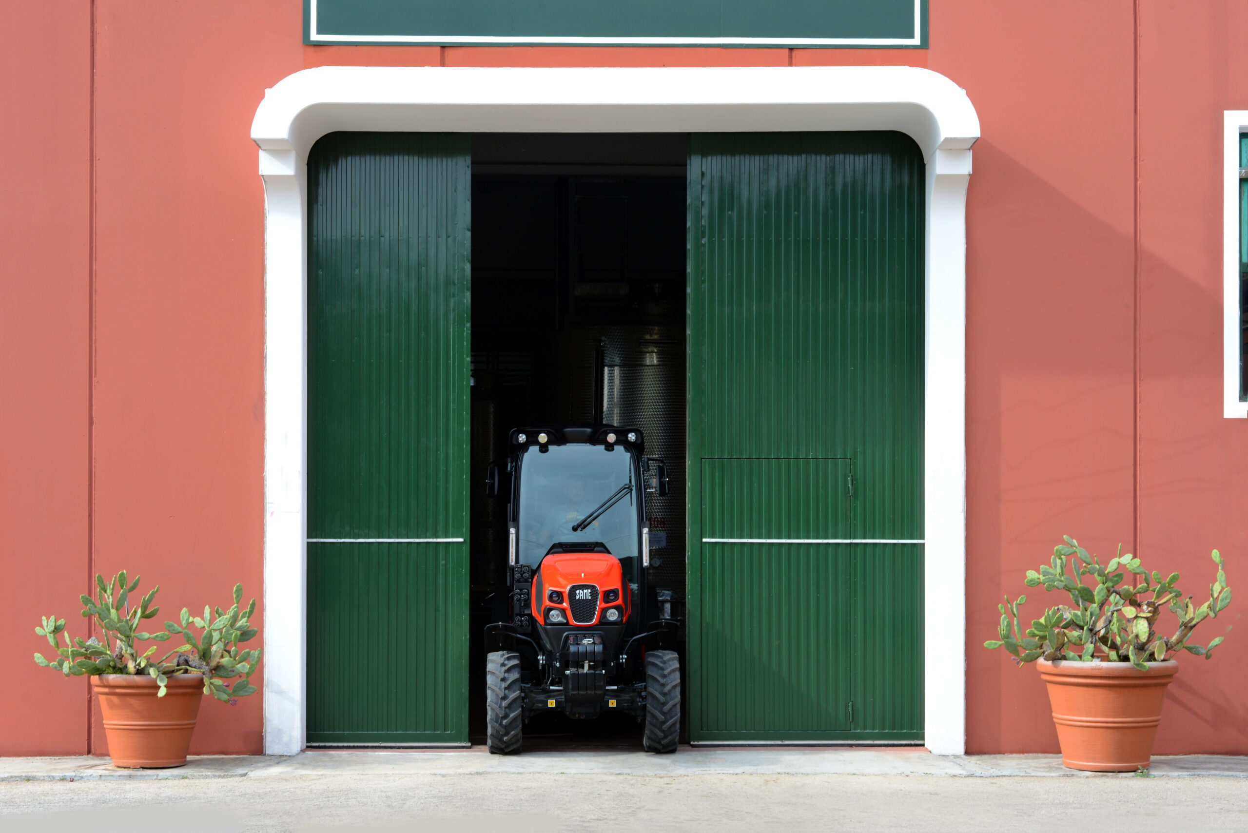 Trattore rosso che esce da rimessa con porte verdi e struttura color mattone