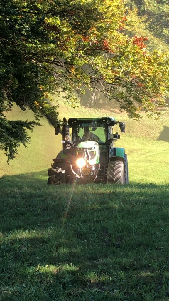 Green agricultural tractor working in a sunny field under trees with headlights shining brightly.
